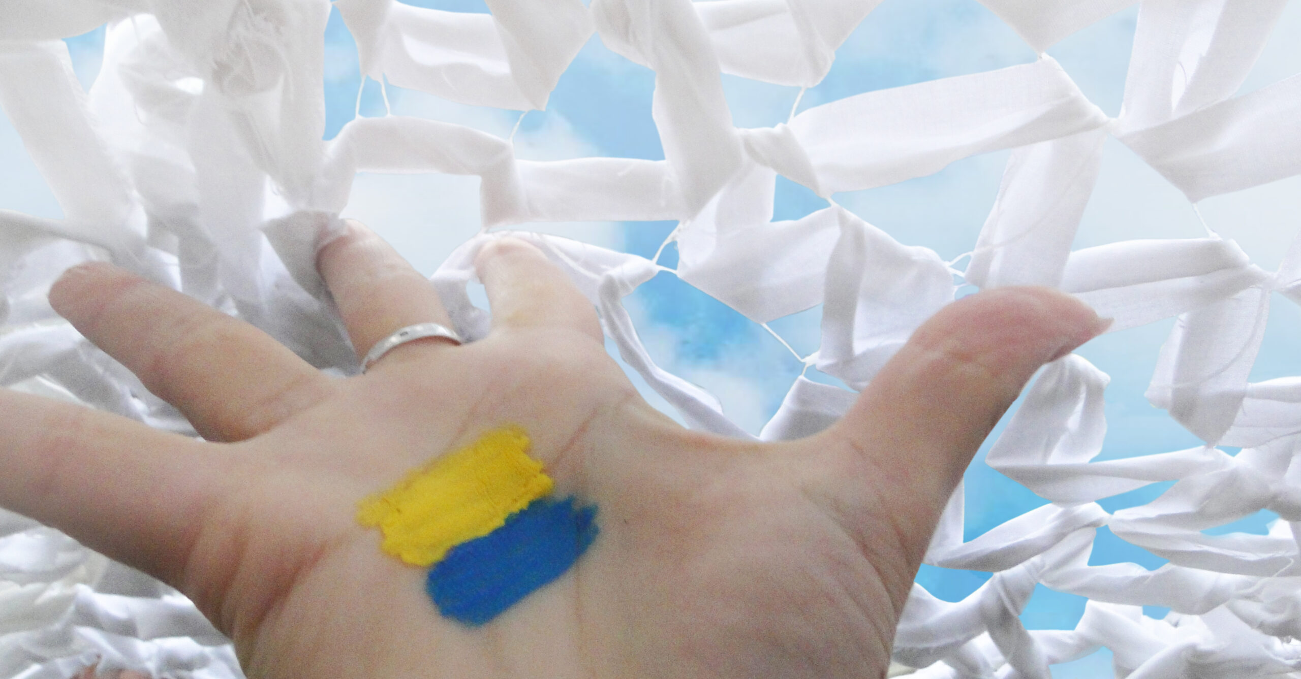 Image of the Ukrainian flag painted on the inside of a hand. The hand is trying to get through a barrier made of white cloth. Image was featured in the Safety Net exhibit.