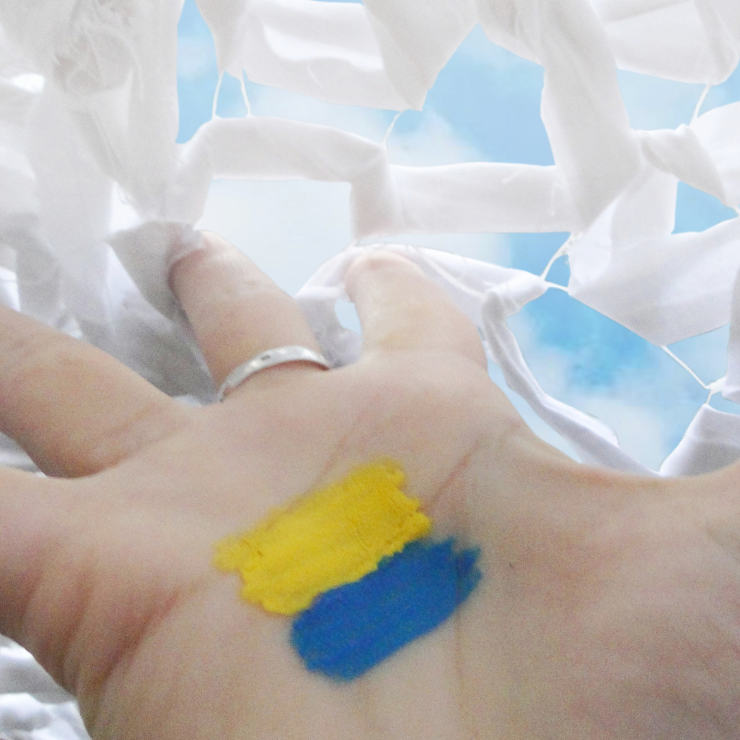 Image of the Ukrainian flag painted on the inside of a hand. The hand is trying to get through a barrier made of white cloth. Image was featured in the Safety Net exhibit.